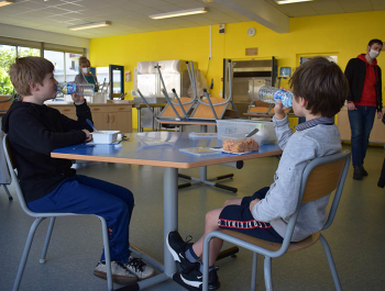 Deux garçons d'une dizaine d'année sont à table dans un réfectoire au premier plan. En arrière-plan, le self et des adultes qui portent des masques.